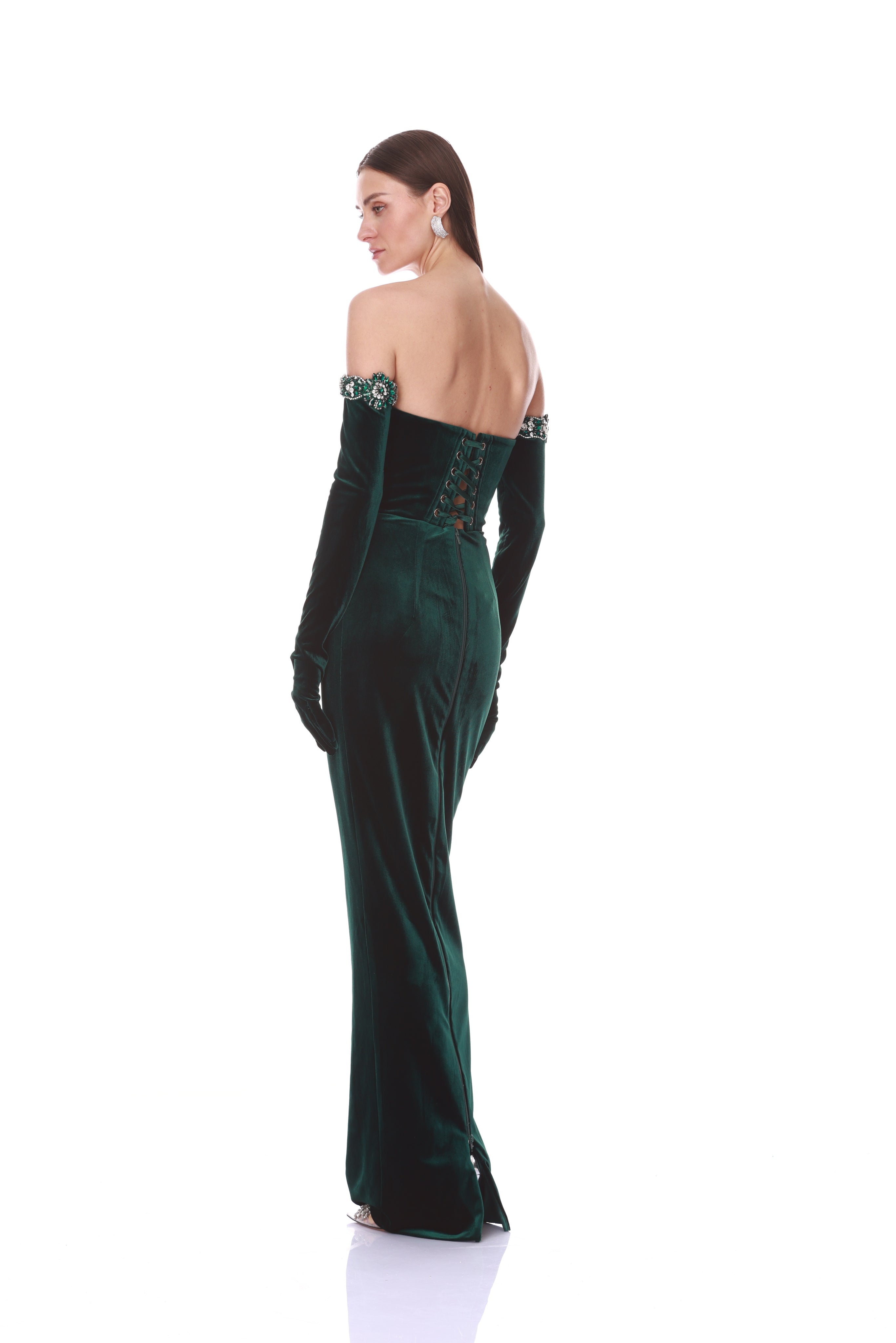 A woman modeling a dark green velvet off-the-shoulder gown with long gloves, shown from the back against a solid white background.