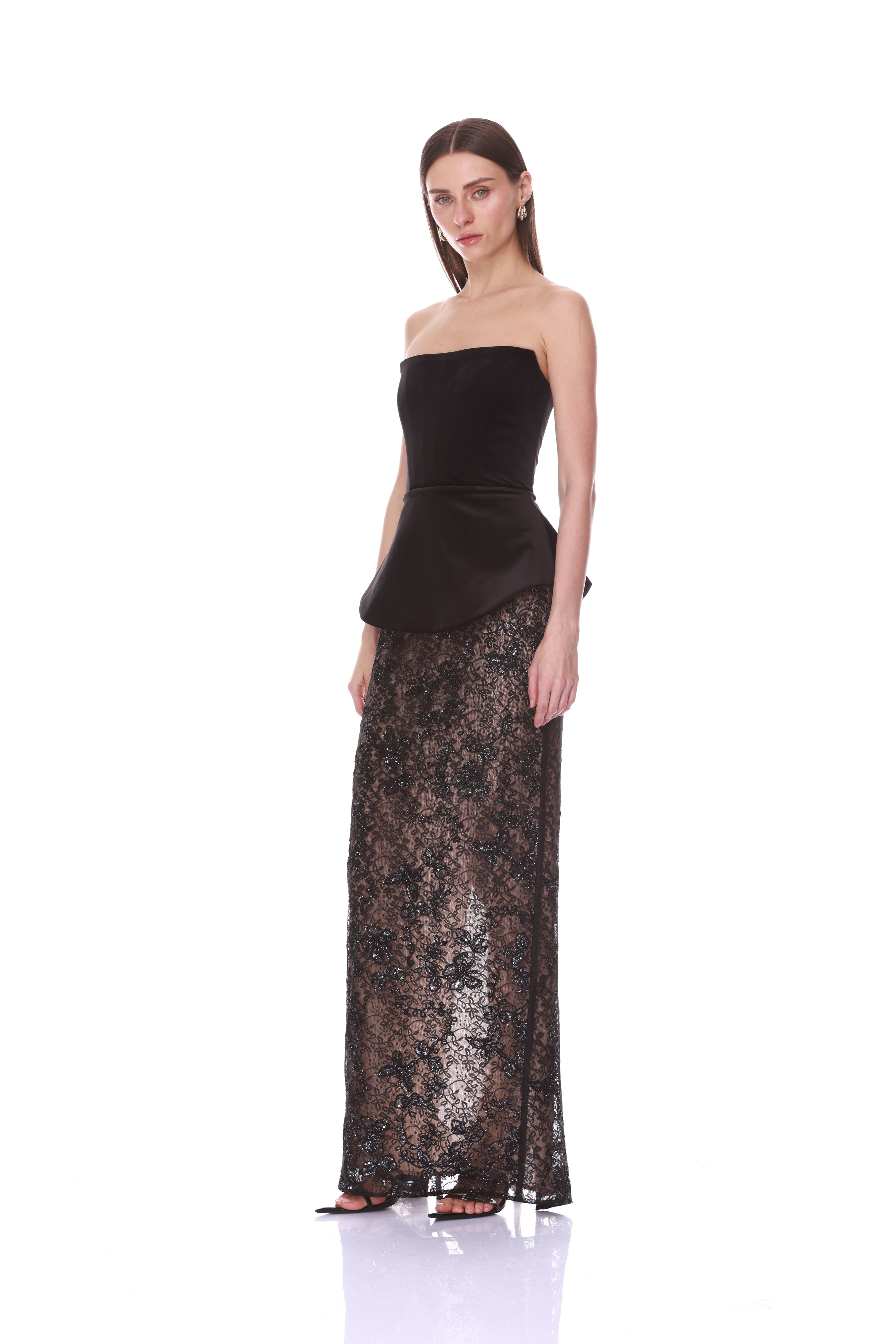 A woman modeling a strapless black velvet peplum top paired with a floor-length sheer black lace skirt, photographed against a seamless white studio background.