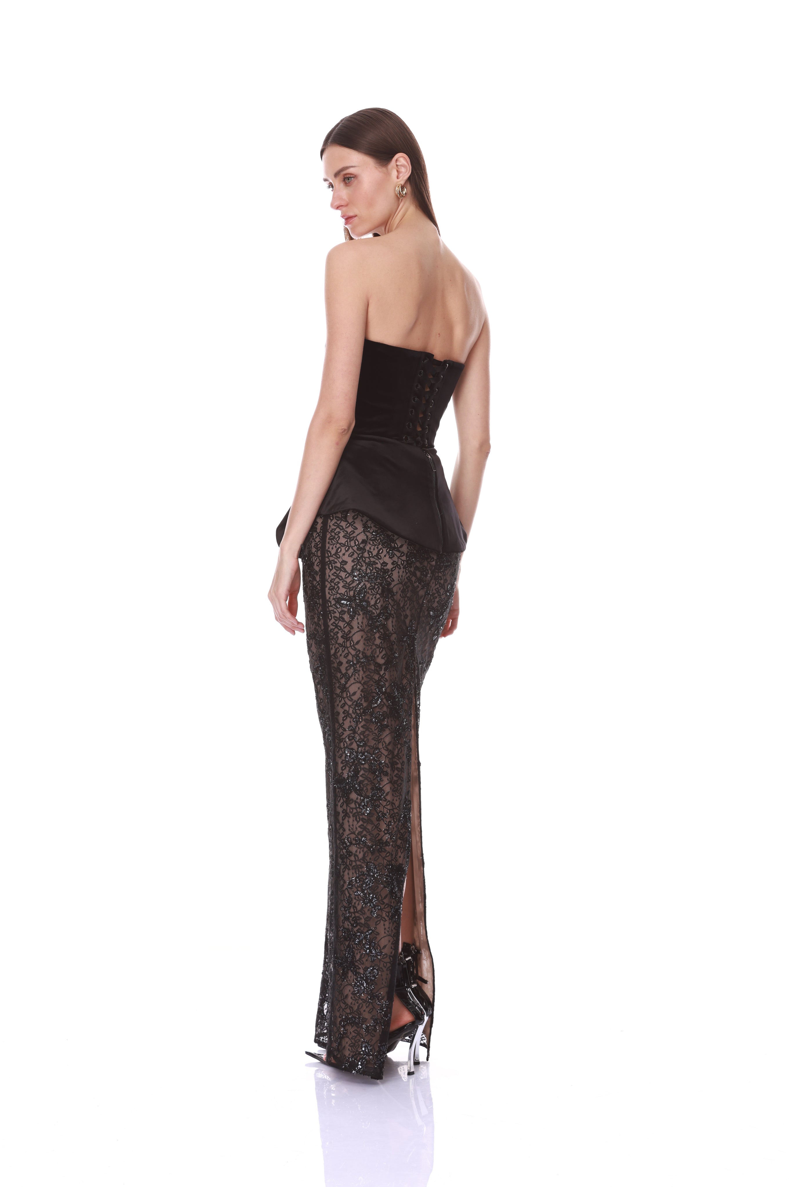 A woman modeling a black strapless lace gown with a structured corset-style bodice and a high slit, photographed from the back against a plain white studio background.