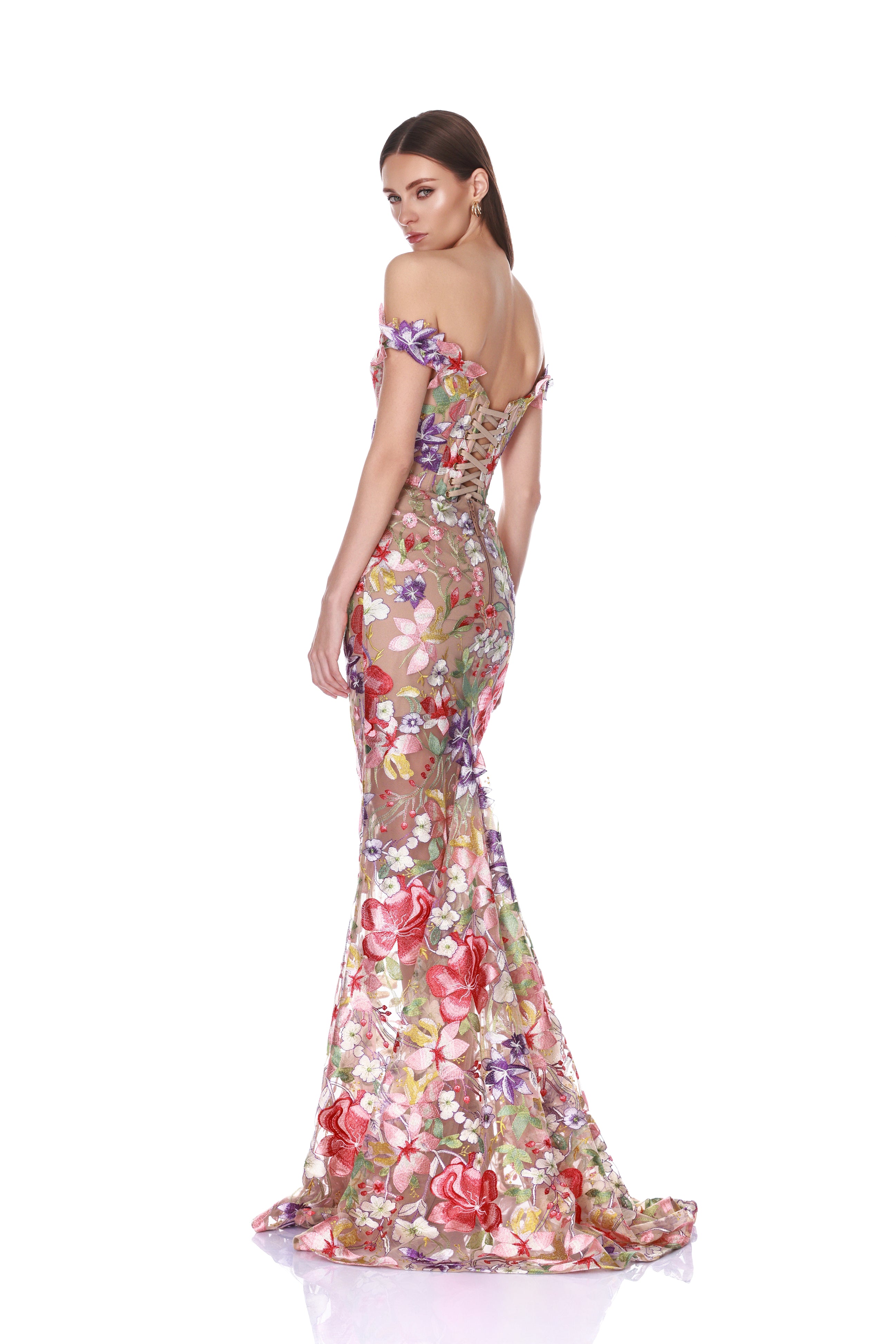A woman modeling a floor-length off-the-shoulder evening gown with a fitted silhouette and dramatic mermaid flare, covered in a colorful floral print on sheer mesh. She is photographed from the back against a plain white studio background.
