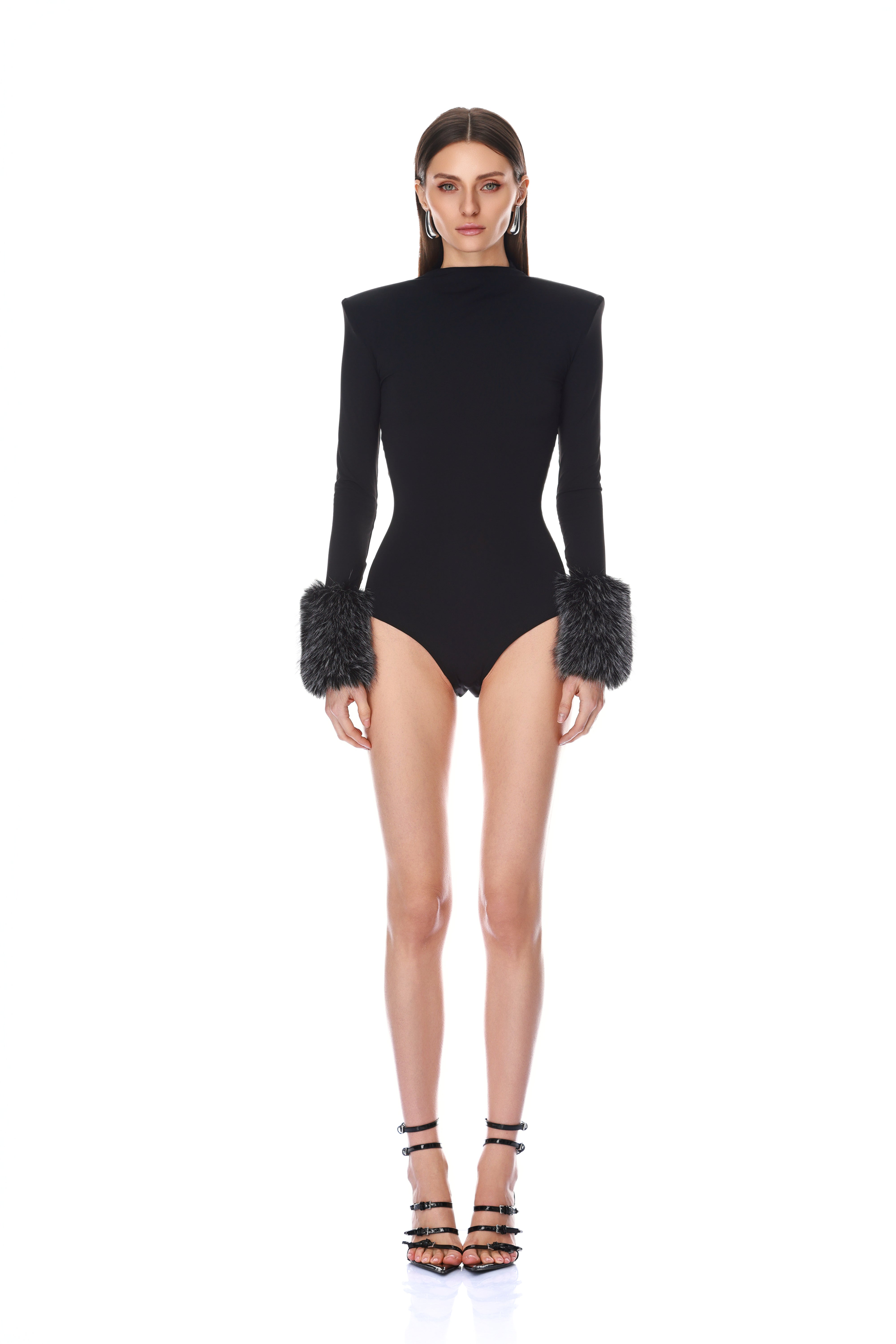 A model stands against a white studio background wearing a black long-sleeve bodysuit with dramatic shoulder pads and dark gray faux-fur cuffs, paired with black strappy high-heeled sandals.